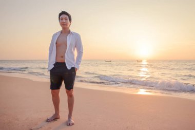 Sahilde kameraya bakarak mutlu bir Asyalı genç yaz tatilinde gün batımında arka planda geziyor..