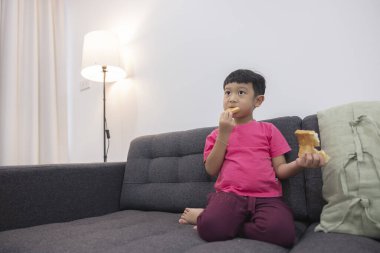 Oturma odasındaki rahat kanepede ekmek yiyip televizyon izleyen gülümseyen küçük çocuk. Dijital akıllı televizyonda oyun oynamaktan zevk alıyorum. Tatlı kullanan küçük mutlu çocuk..