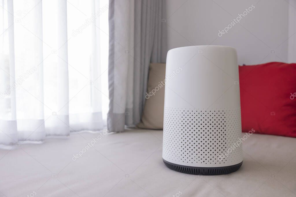 White air purifier sits in the living room with white curtains and red-brown pillows in the background. Protect PM 2.5 dust and air pollution concept. air cleaner removing fine dust in house.