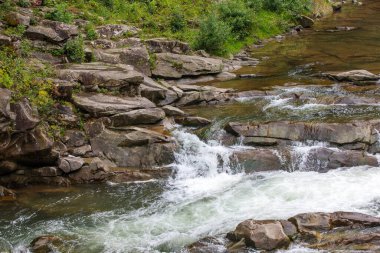 Hızlı bir dağ nehri, daha küçük taşlar ve çakıl taşlarıyla çevrili düz kayaların üzerinden akar. Su gözle görülür şekilde çalkantılı, kayaların üzerinde hızla hareket ederken beyaz köpük oluşturuyor..