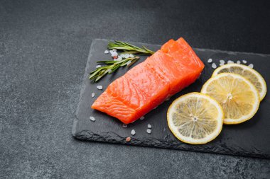 Taze somon filetosu yanında koyu bir yüzey üzerinde limon dilimleri ve otlar..