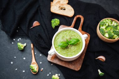 Kremalı yeşil brokoli çorbası fesleğen garnitürlü bir kâsede sunuluyor, yanında ekmek dilimleri ve koyu renk kumaş üzerinde sarımsak.