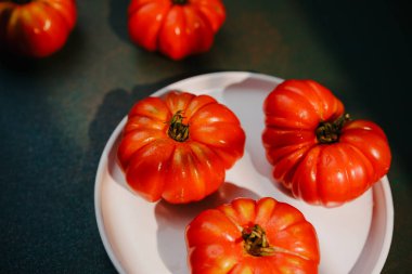 Ripe red heirloom tomatoes sit on a simple white plate with a dark textured background.