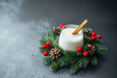 Glass of creamy winter drink with cinnamon stick on festive pine branch and red berries