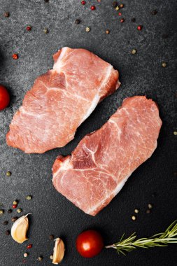 İki domuz pirzolası karanlık bir yüzeye yerleştirilir ve etraflarında otlar ve sebzeler vardır..