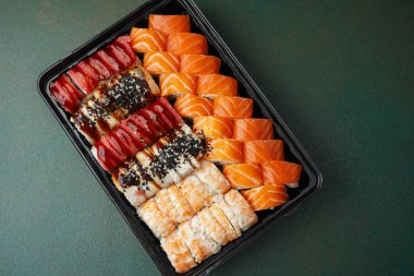 Suşi, yeşil arka planda siyah tepside somon balıklı karides yılan balığı çeşitleri yuvarlıyor.