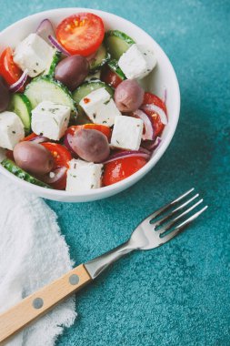 Beyaz kâsede beyaz peynirli Yunan salatası ve mavi arka planda çatal.