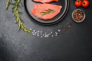 Siyah seramik tabakta çiğ biftek, domates ve mutfak masasında baharat.