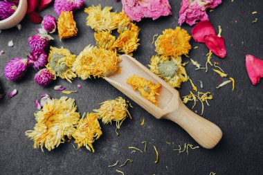 Kurumuş sarı ve pembe çiçekler, ahşap bir kepçeyle koyu dokulu bir yüzeye saçılmış.