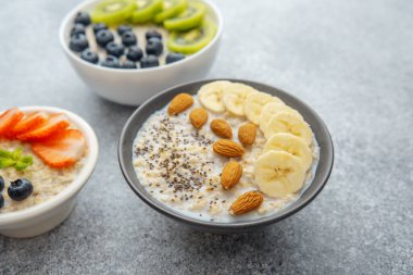 Üç yulaf ezmesi kasesi ve gri arka planda kivili muz ve çilek.