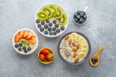 Üç yulaf ezmesi kasesi ve gri arka planda kivili muz ve çilek.