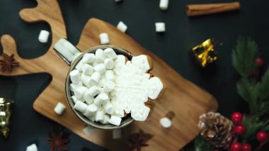 Şekerlemelerle dolu sıcak bir kupa ve tahtada kar tanesi kurabiyesi, etrafta bayram süsleri.