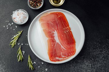 Beyaz tabakta dilimlenmiş jambon ve siyah arka planda baharatlar.