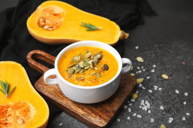 Tohumlar ve otlarla kaplı bir kase kremalı kabak çorbası. Koyu bir sayfada tereyağlı kabak yarısıyla servis ediliyor.