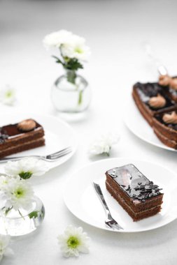 Beyaz çiçekli zengin çikolatalı pastalar ve temiz bir masada beyaz tabaklarda gümüş çatallar.