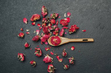 Küçük kurumuş gül tomurcukları ve yaprakları, elinde tomurcuklar olan bir tahta kaşıkla karanlık bir yüzeye saçılmış..