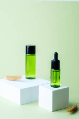 Kurumuş çimen aksanıyla süslenmiş beyaz kaideye siyah kapaklı yeşil cam damlalar ve şişe.