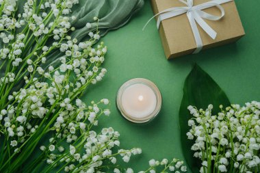 Vadideki zambaklarla çevrili yanan mum ve yeşil arka planda paketlenmiş hediye.