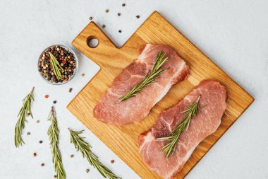 İki çiğ domuz pirzolası üzerinde taze biberiye filizleri olan bir kesim tahtasının üzerinde oturur..