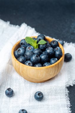 Ahşap bir kase, kumaş üzerinde nane yaprağı olan dolgun yaban mersinleri taşır..