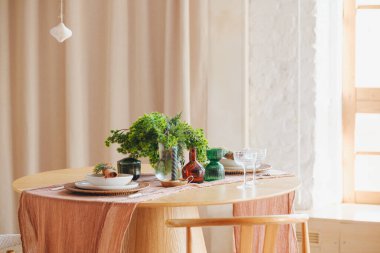 Vazoda yeşil yapraklar, dekoratif şişeler ve yer dekorasyonu ile yuvarlak yemek masası.