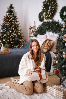 Hediye kutusunu tutan genç bir kadın süslü Noel ağacının yanında rahat bir odada.
