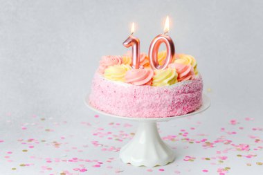 Üzerinde mumlar olan pembe bir doğum günü pastası konfetiyle çevrili beyaz bir kaidede duruyor..