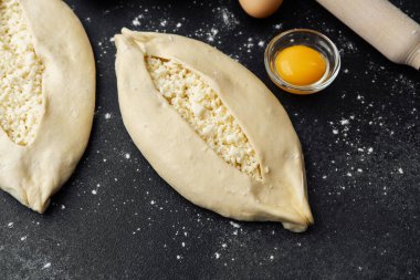 İki çiğ khachapuri ekmeği, içinde fırın malzemeleri olan siyah un kaplı bir masada.