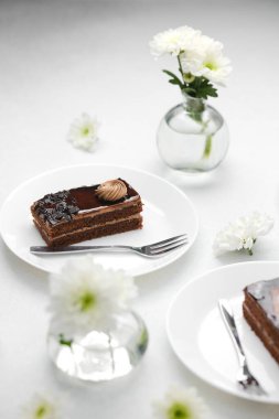 Vazo sahnesinde beyaz çiçeklerin yanında çatalla çikolatalı pasta dilimi.