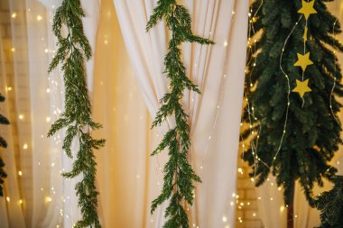 Yeşil çam çelenkleri ve yıldız süslemeleri bir perdenin yanındaki Noel ağacında asılı..