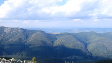 Koyu yeşil dağ sıraları bulutların dağıldığı parlak bir gökyüzünün altında sisli bir ufka doğru uzanıyor..