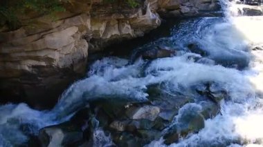 Koyu kayalıkların üzerinde dalgalı beyaz su taşar. Tepesinde yeşil yapraklar vardır.