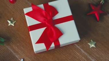 Kırmızı kurdeleli beyaz hediye kutusu ve ahşap masa arkasında şenlikli süsler..