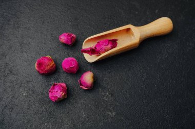 Kurumuş pembe gül tomurcukları, tek bir tomurcuğu tutan ahşap kepçeyle karanlık bir yüzeye saçılmış..