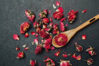 Kurumuş kırmızı gül tomurcukları ve yaprakları bir tomurcuğu tutan tahta bir kaşıkla karanlık bir yüzeye saçılmış..