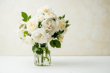 Beyaz masadaki cam vazoda bej desenli duvar arkaplanı olan beyaz gül buketi.