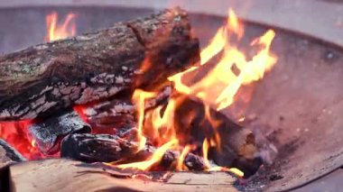 Parlak turuncu alevlerle yanan odunlar ve açık havada yanan metal bir ateş çukurunun içinde parlayan kırmızı közler..