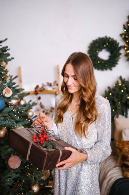 Payet elbiseli genç bir kadın Noel ağacının yanında bir hediye kutusu tutuyor.