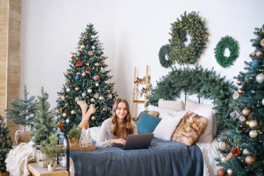 Yatakta dizüstü bilgisayar kullanan kadın süslü Noel ağaçları ve şenlikli çelenklerle çevrili.
