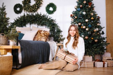 Noel hediyesini elinde tutan kadın süslü ağaç ve yatağın yanında oturmuş bayram süslemeleriyle.