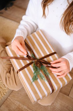 Çizgili hediye kutusuna çam filizi ve koni detaylarıyla kurdele bağlayan eller..