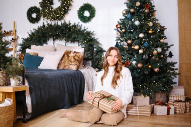 Kadının elinde Noel çelengi ve ışıklarıyla süslenmiş ağaç ve yatağın yanında bir hediye var.