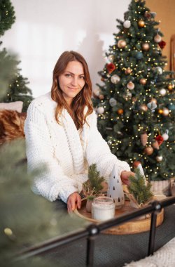 Süslü Noel ağacının yanında gülümseyen, tepside şenlikli bir dekor tutan rahat kazaklı bir kadın.
