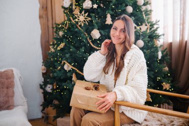 Samimi süveter giyen gülümseyen kadın hediye kutusunu kapalı bir yerde, Noel ağacının yanında tutuyor.