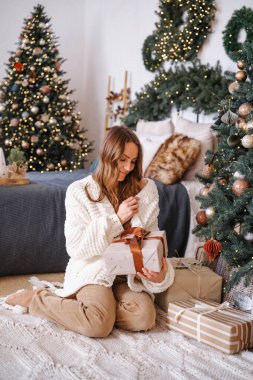 Noel hediyesi kutusu tutan bir kadın, süslü bir ağacın yanında, rahat bir halının üzerinde, sıcak bir atmosferde.