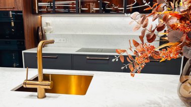 Zarif mutfak içi altın musluk, lavabo, beyaz tezgah, koyu dolaplar, sonbahar yaprakları.