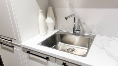 Parlak paslanmaz lavabosu, krom musluğu, beyaz mermer tezgahı, beyaz dolapları, vazoları olan modern bir mutfak..