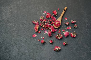 Kurumuş kırmızı gül tomurcukları ve taç yaprakları, tahtadan kaşıkla karanlık yüzeye saçılmış.