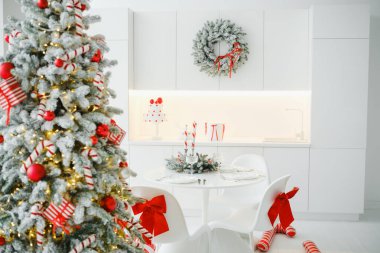 Parlak mutfak, şenlikli Noel ağacı, yemek masası ve kırmızı kurdelelerle süslenmiş sandalyeler.