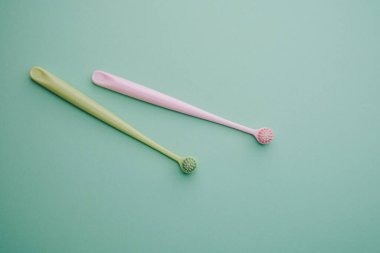 İki pastel renk dil temizleyicisi, pembe ve yeşil, sade bir deniz mavisi zemininde uzanıyor..